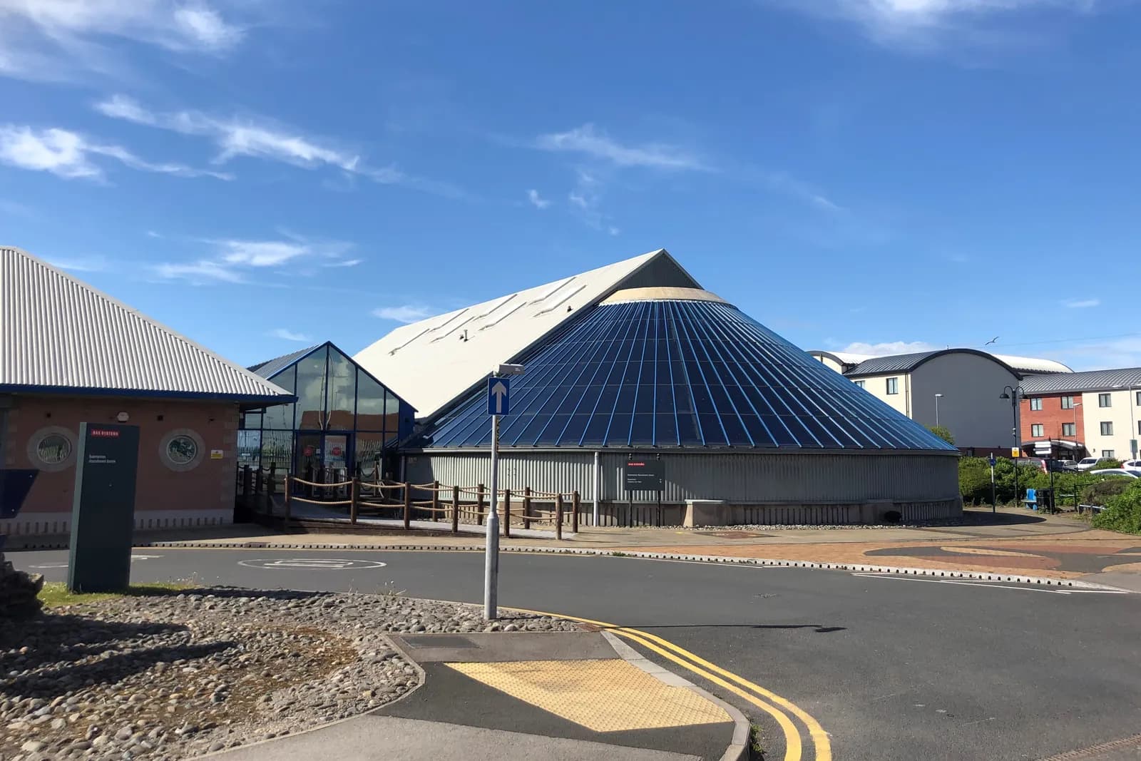 A modern building with a large conical glass roof and a pitched white metal roof under a bright blue sky with wispy clouds.